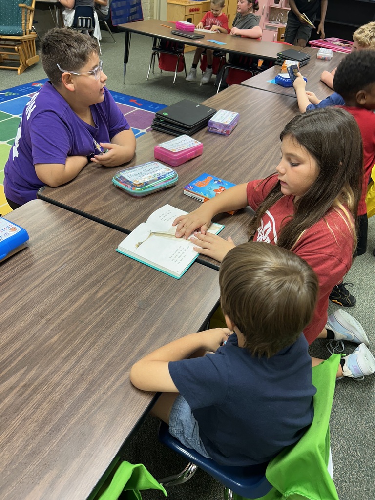 A classroom with children seated at tables. Two children read books and interact with a teacher. Tables have books and bags.