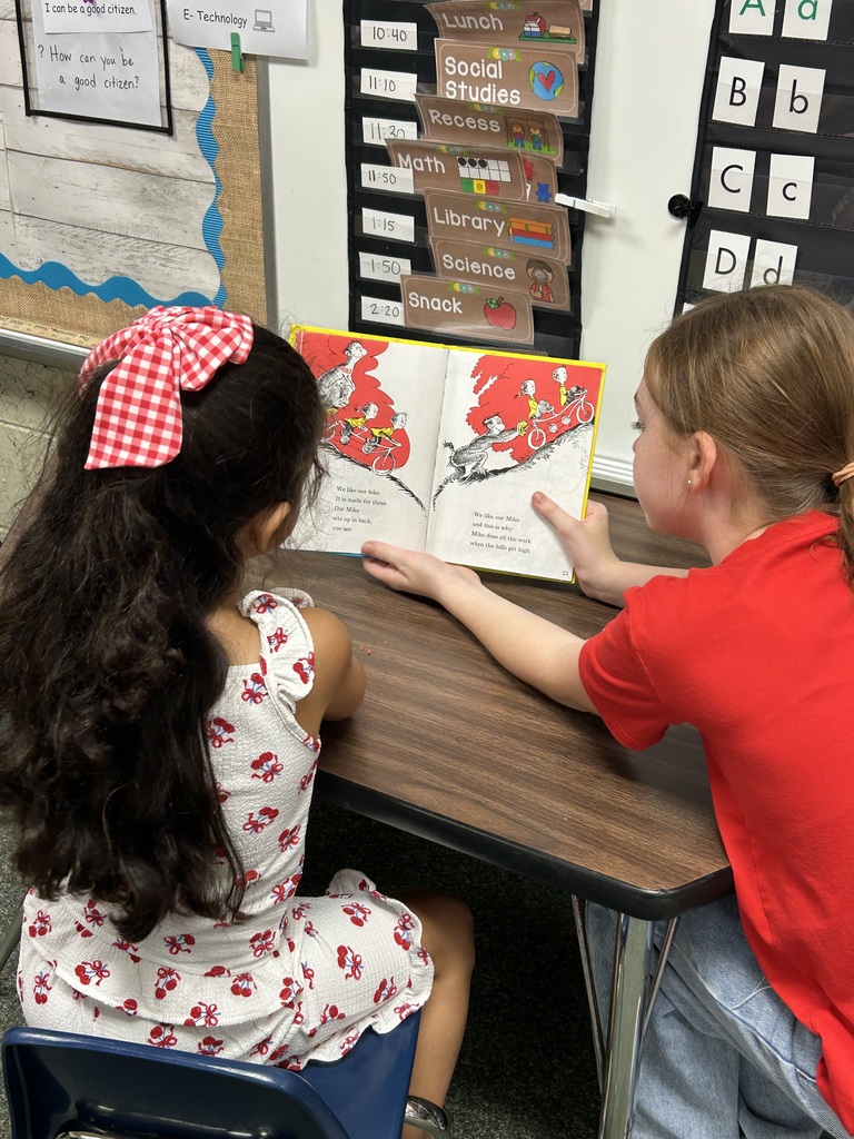 Two girls seated at a table, one in a white dress and the other in red, reading a book together.
