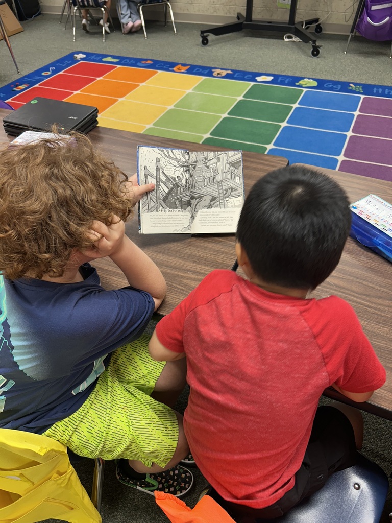 Two children, one in blue and the other in red, are seated on chairs at a desk, looking at a book.