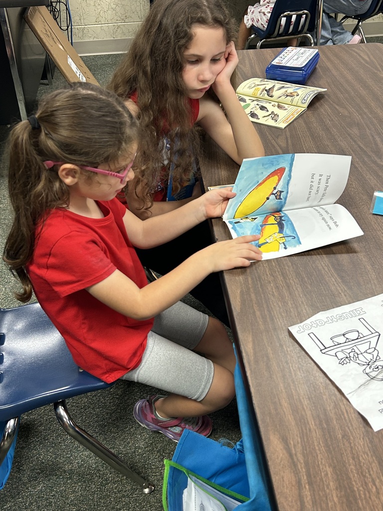 Two girls sit at a table, one reading a book with pictures, and the other studying a book with text.