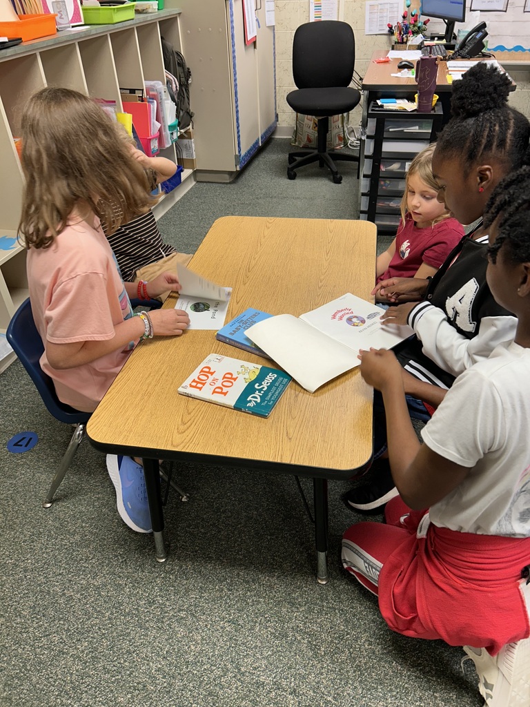 Five children in a classroom sit at a small table. They each have a book in front of them.