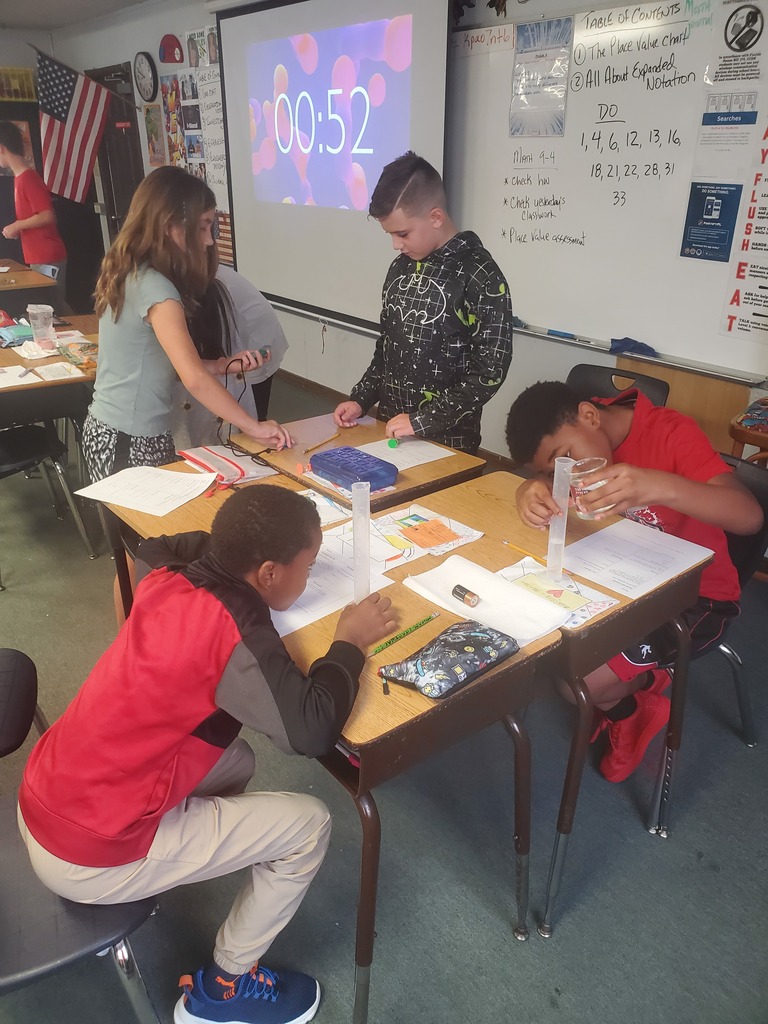 A classroom with four children seated at a table, working on a project. Papers and materials are on the table. A projector screen shows a timer.