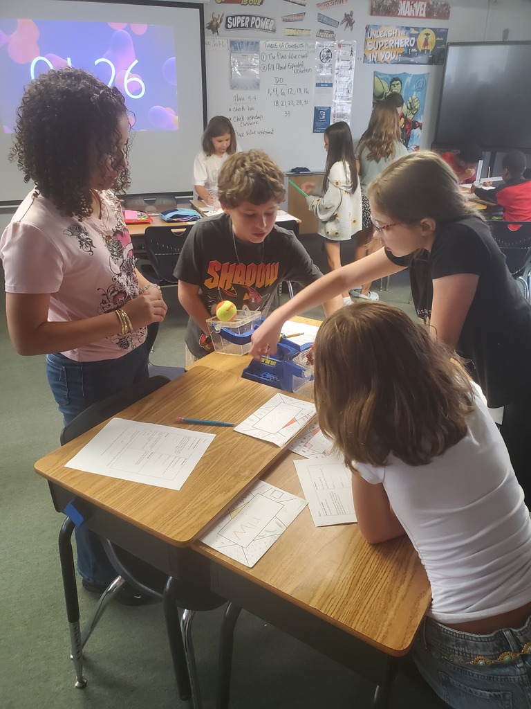 Five children in a classroom are working around a table with papers and a toy. One child holds a toy while others look at the papers.