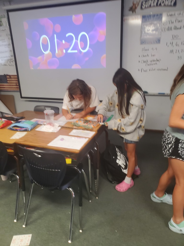 Two individuals in a classroom, one sitting at a desk and the other standing. A projector displays a timer.