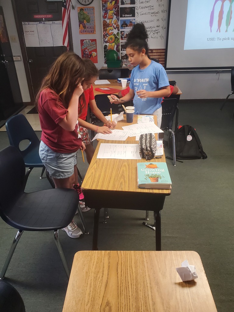 Three children work at a desk, two standing, one seated, with papers and a book. A projector screen displays behind them.