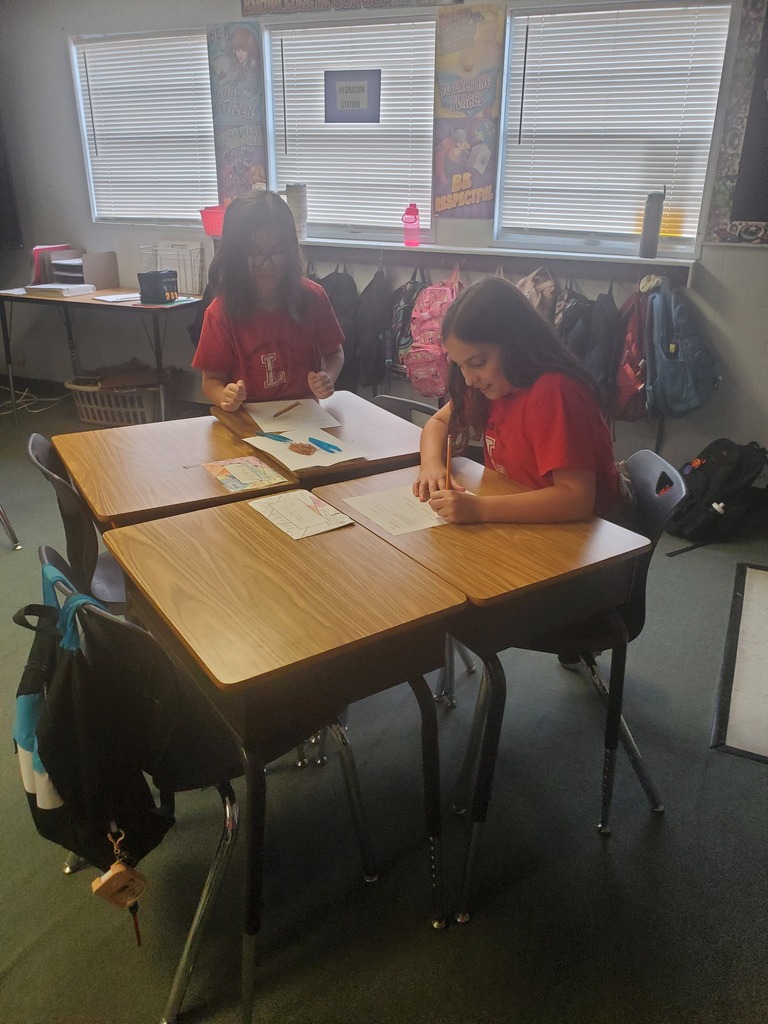 Two students seated at desks in a classroom, each writing on paper. Windows and backpacks are in the background.