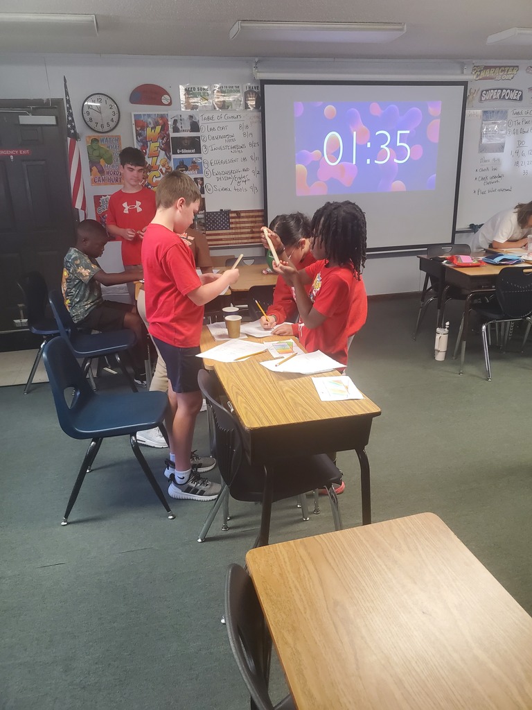 A classroom with students. Some sit at desks while others stand. One student uses a phone. A projector displays a timer.