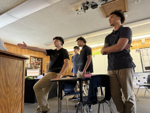 Three males stand in a classroom. One gestures with arms extended. Two stand with arms crossed.