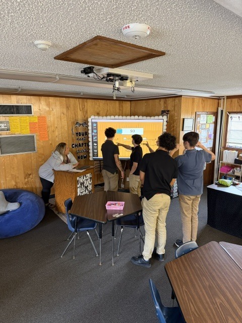 A group of people stand around a table in a classroom with a projector screen, wooden walls, and a white ceiling.