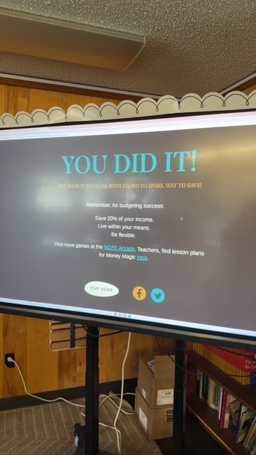 A computer screen displays the message "You Did It!" in large letters. Below, there are budgeting tips and a mention of games. Wooden walls and a shelf with books surround the screen.