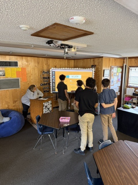 A group of people stand around a table, facing a screen that displays a presentation. The room has wooden walls and carpeted floor.