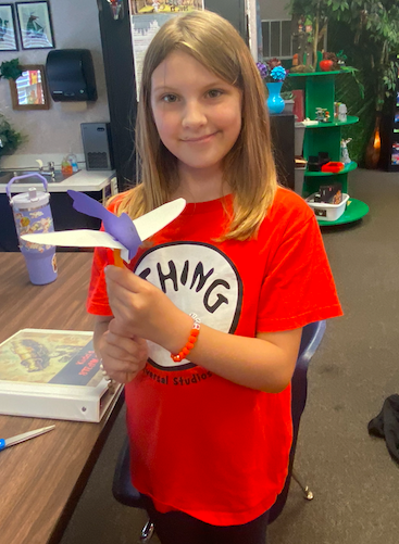 A girl in a red shirt holds a purple and white paper airplane, standing in a classroom with a desk and books in the background.
