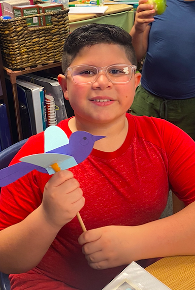 A young boy with glasses holds a blue and white paper bird in a classroom with shelves in the background.