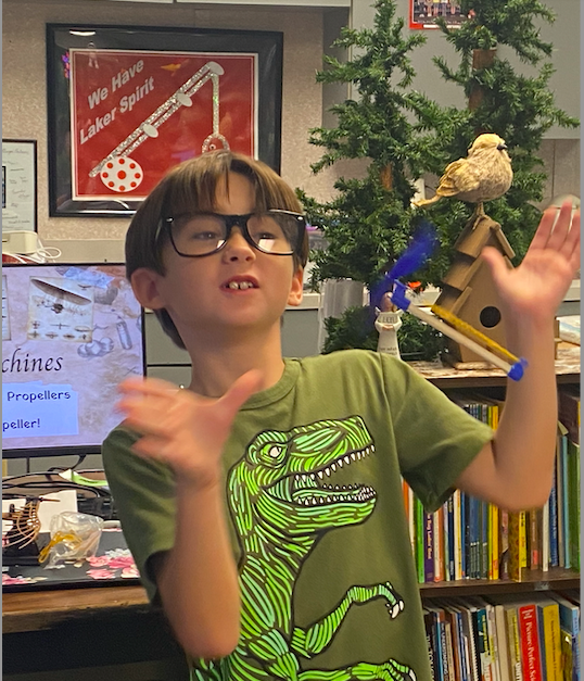 Boy with glasses and dinosaur shirt stands in front of a bookshelf. He is animatedly gesturing with his hands.