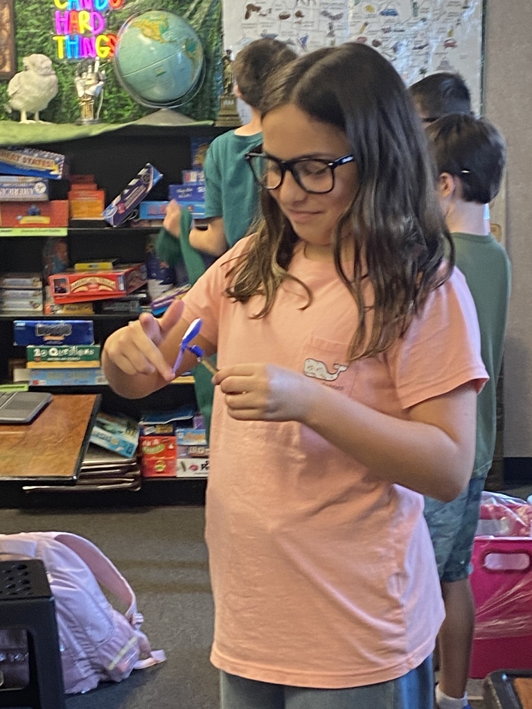 A girl with glasses holds a blue object in a classroom with a globe and books.