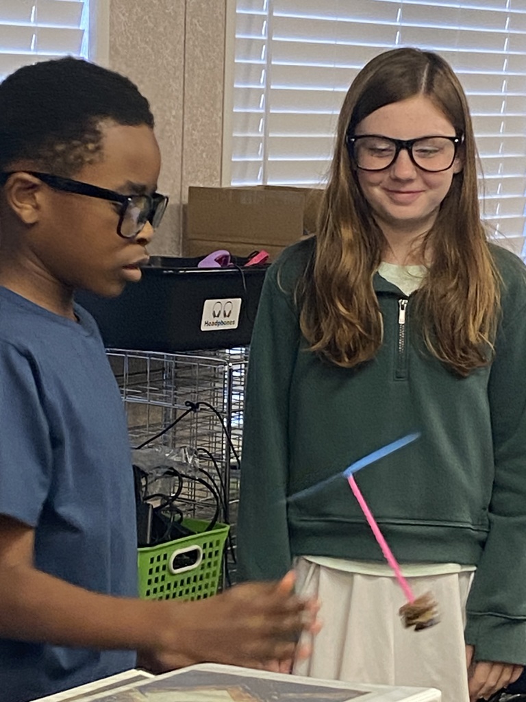 Two students in a classroom, one holding a toy and the other looking at it, with storage boxes and a cage behind them.