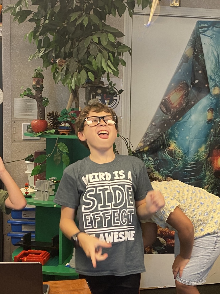 A boy wearing glasses and a yellow shirt bends over next to a tree in a room.