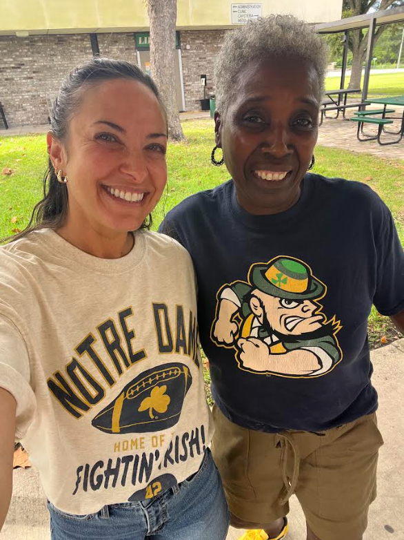 Two women smile at the camera. One wears a white Notre Dame shirt, the other a blue shirt with a leprechaun. A tree and building are in the background.