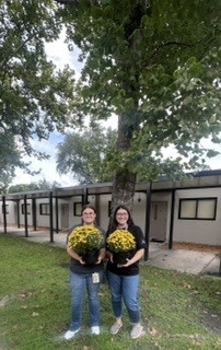 Photo of two women holding yellow mum flowers in front of a tan building and large tree.