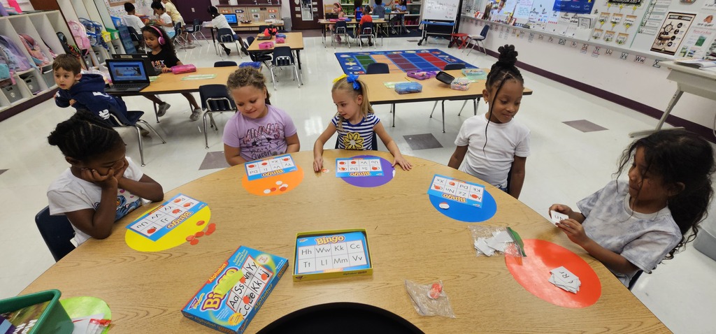 Five young children sit around a table, engaged in an activity with colorful cards. Background includes other tables and children.