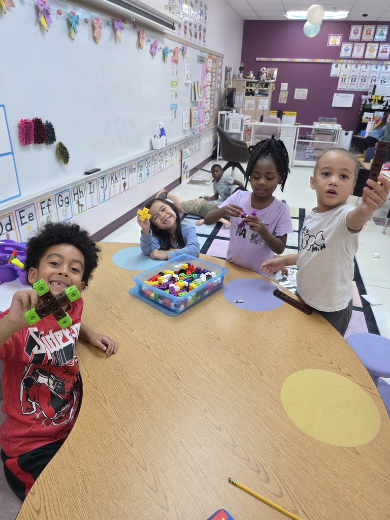Four children seated at a table in a classroom. One holds a colorful structure and another holds up a phone.