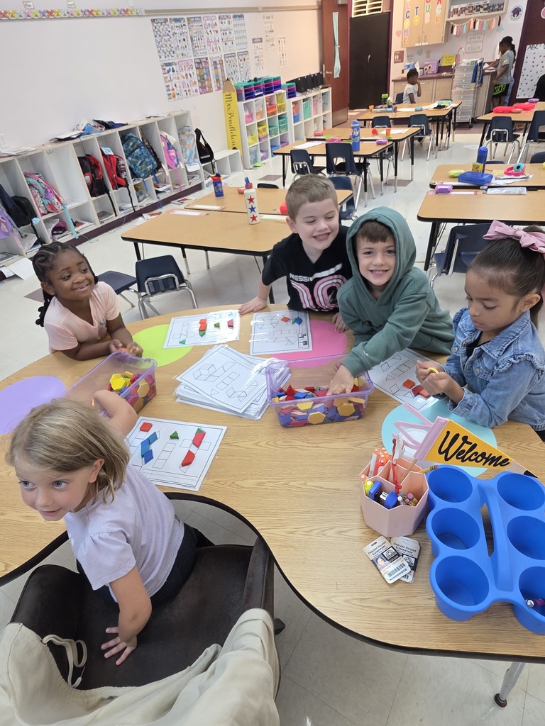 Children in a classroom are seated at a table with educational materials, including papers and colored markers.
