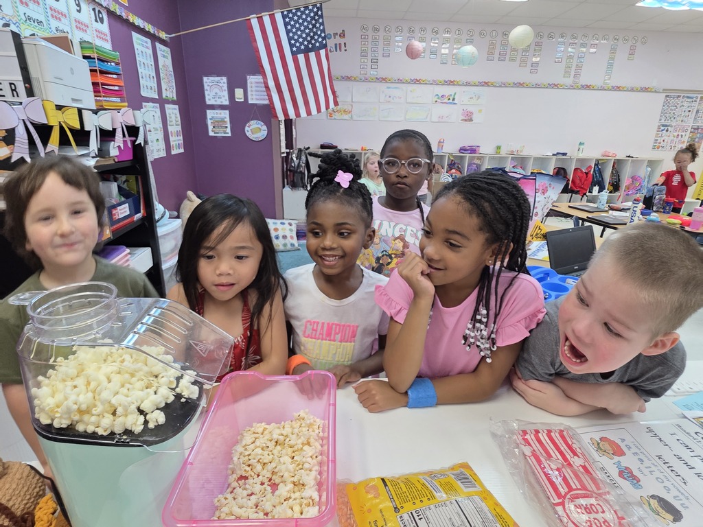 Children sit at a table with popcorn in a classroom. The table has popcorn and other items.