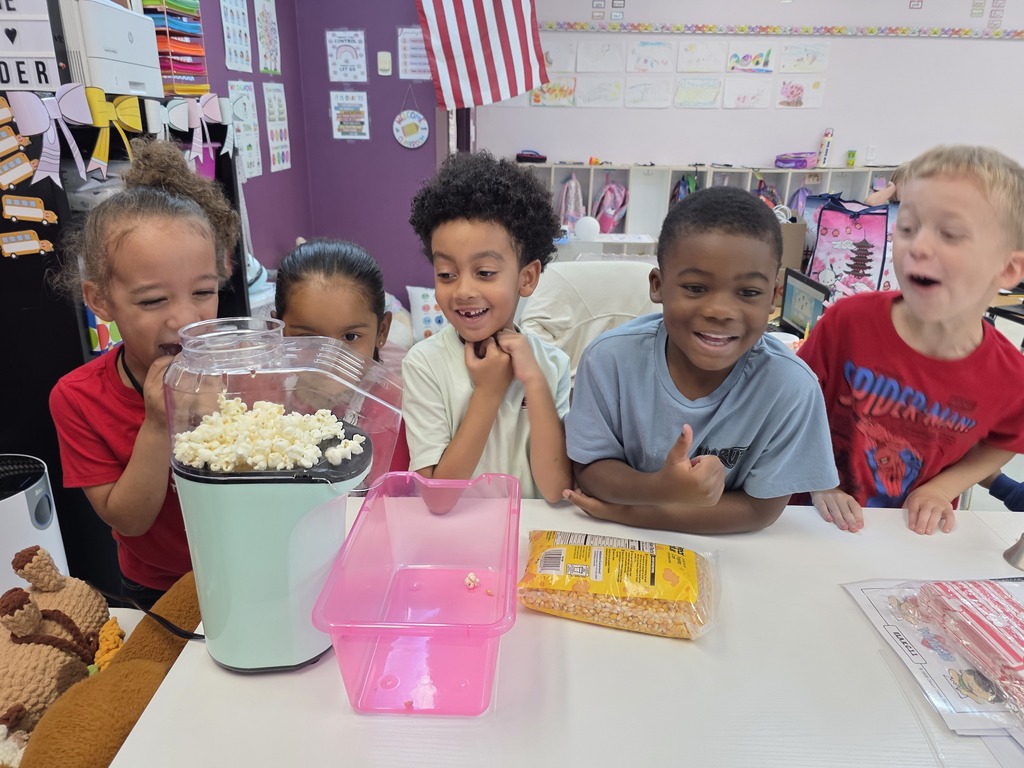 Five children seated around a table, with popcorn in a machine, seated against a backdrop of classroom decorations.