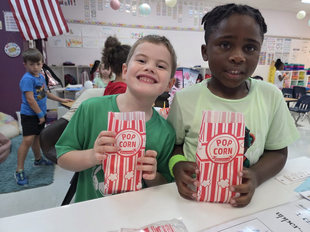Two kids seated at a table holding popcorn boxes. A flag and balloons are in the background.