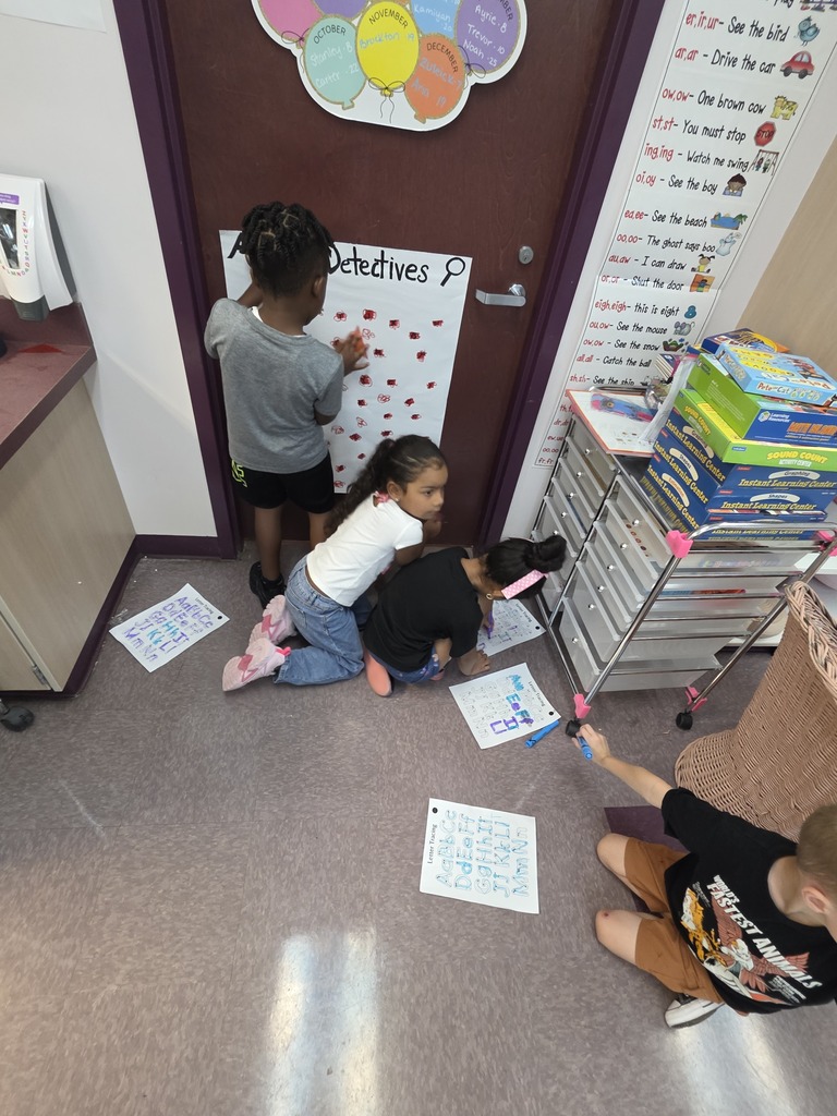 Children work on a board by a door, one holding a marker, others with papers on the floor.