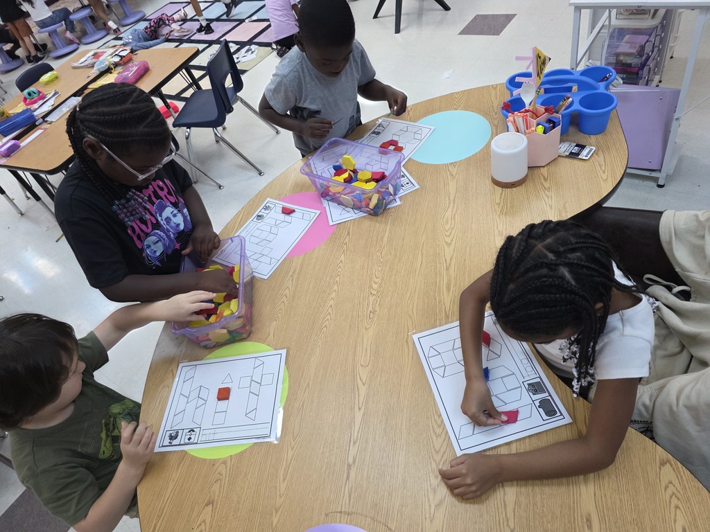 Four children seated at a table with educational materials, including a circular object and a worksheet.