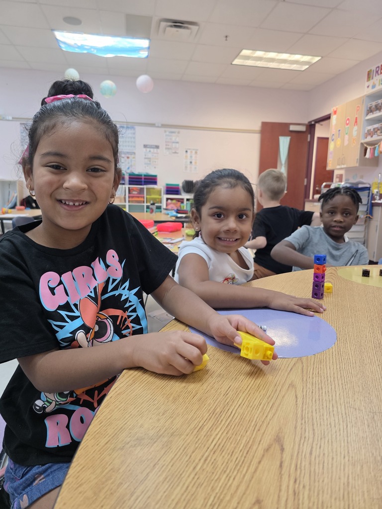 Three children seated at a table, one wearing a black shirt with "Girls" text, smiling. The table has toys. Background includes a room with various items.