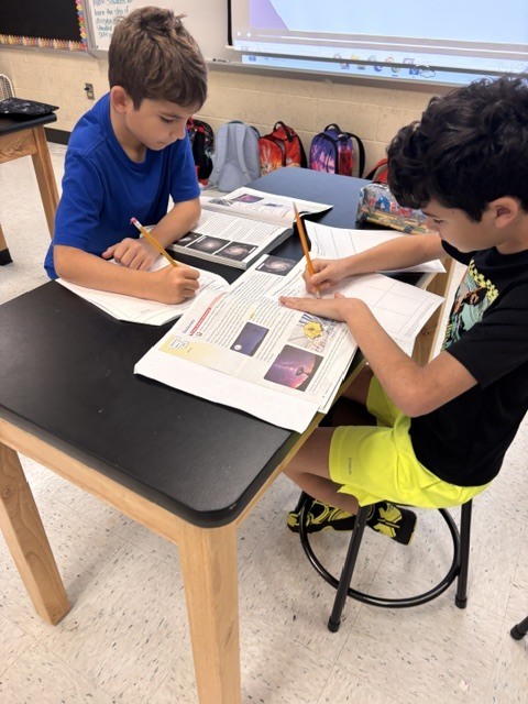 Two boys at a desk, each with a book and pencils, engaged in writing activities.