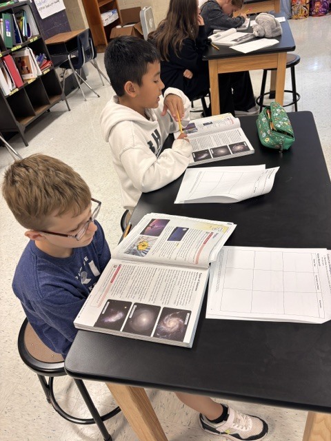 Two children are seated at a table, engaged in reading from open books. One child wears glasses.