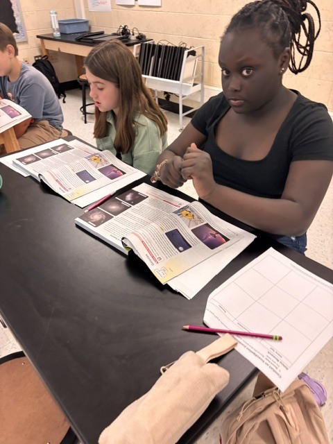Two girls sit at a table, reading and discussing a book. One girl holds a pencil, and a handbag is nearby.