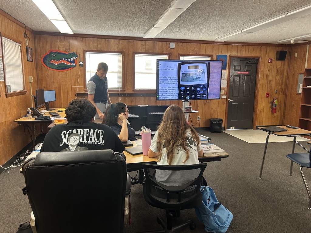A small classroom with a large screen. Four people are seated around a table. A person stands at a window.