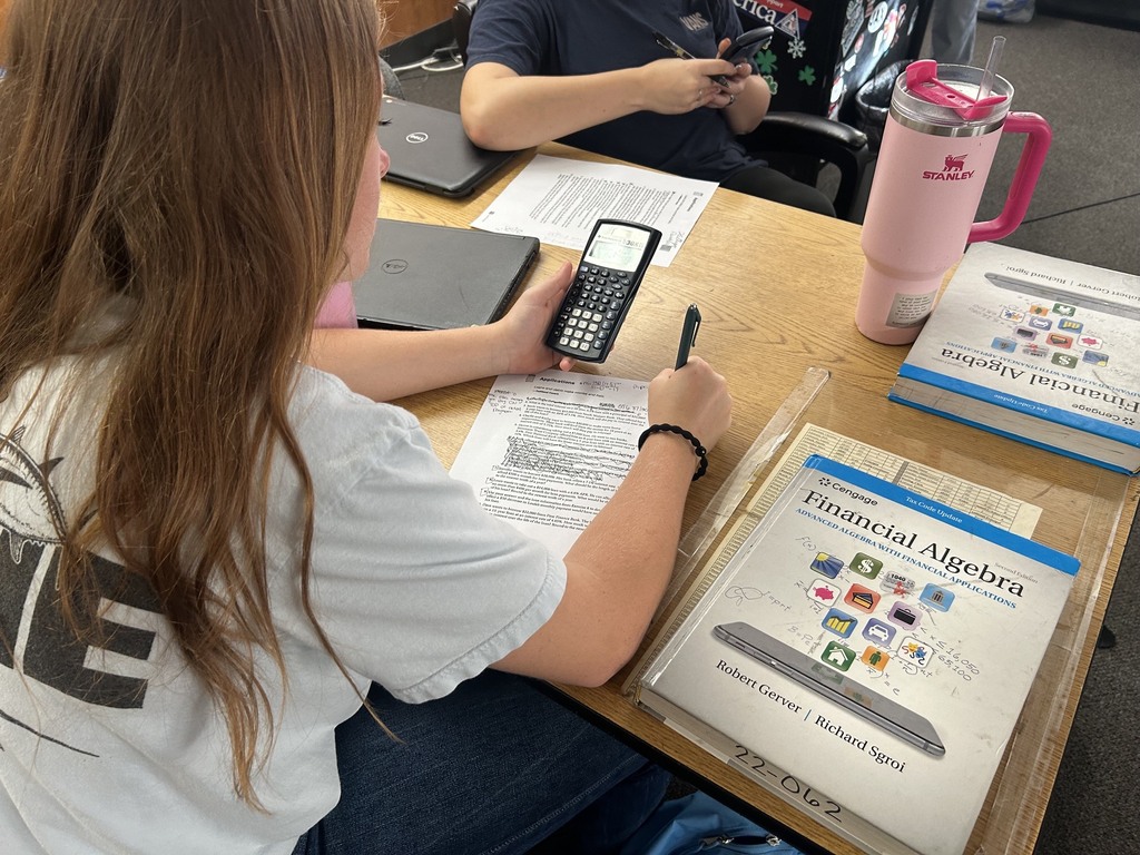 Student seated at a desk, using a calculator and pen to write on paper. Books and a laptop nearby.