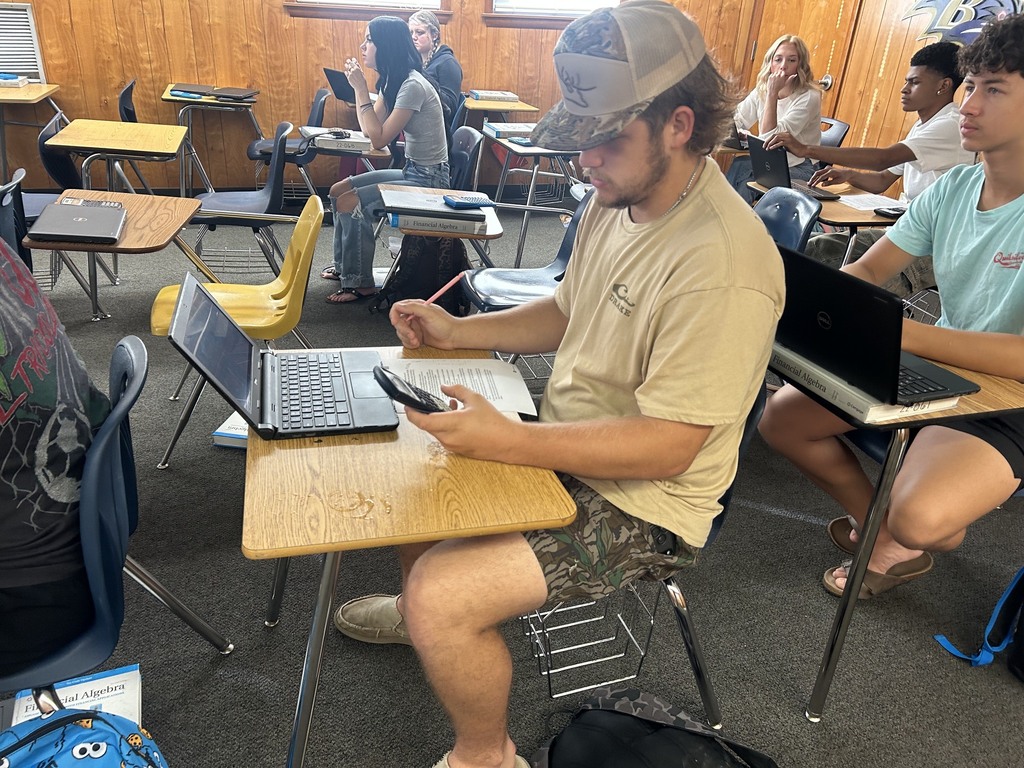 People in a classroom with wood paneled walls, seated at desks with laptops and a person typing on a phone.