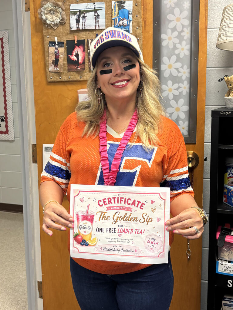 Woman wearing an orange shirt with blue trim and a cap holding a certificate. Behind her is a door with pictures and a shelf with items.