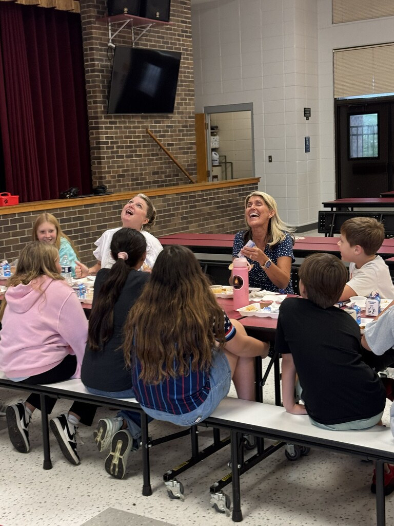 Group of children seated at tables in a cafeteria, a woman stands before them. Behind, brick walls, a stage, and a TV monitor.