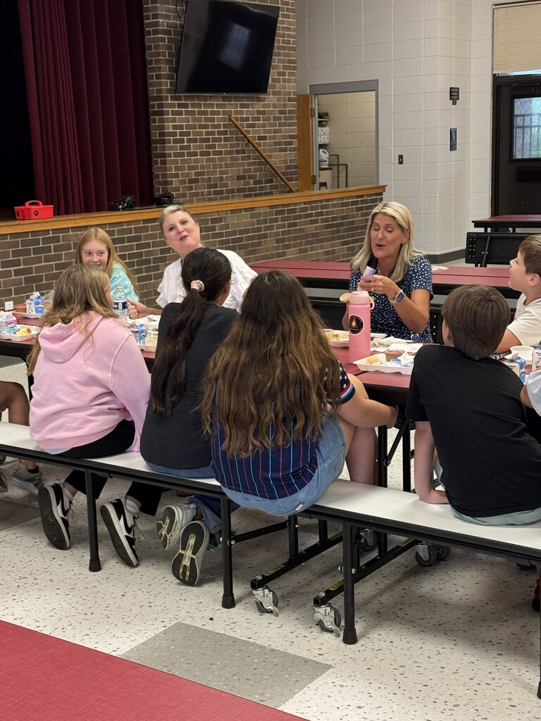 Group of children seated at a table, eating. Behind them is a red curtain, brick wall, and televisions.