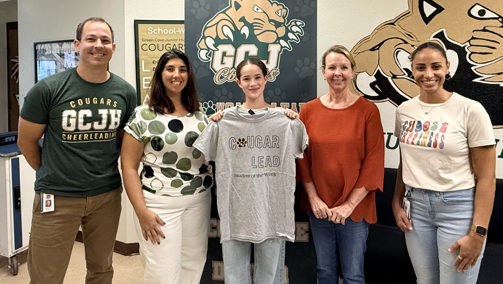 Five adults stand together; one holds a gray t-shirt. Behind them is a banner with a logo and large lion.