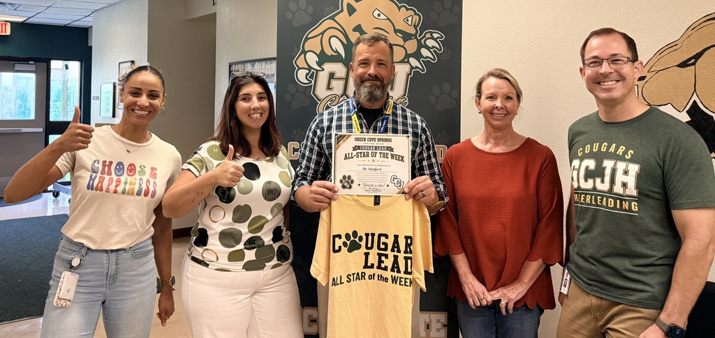 Five adults standing together, holding a certificate and T-shirt. The man in the center wears glasses.