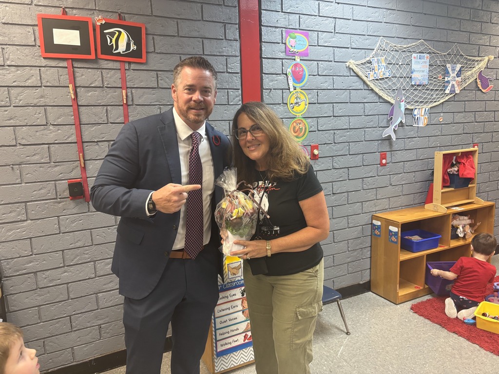 A man and a woman stand together in a classroom, with the man gesturing. The woman holds flowers. Behind them is a brick wall and a play area.