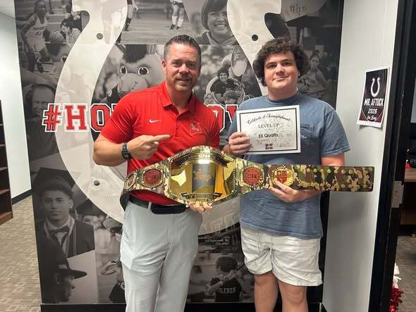 Two men stand with a championship belt; one holds a certificate. Behind them, a mural displays sports history.