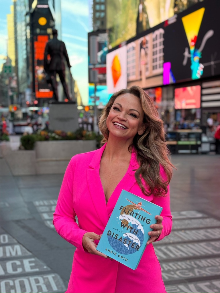 Woman in pink suit stands outdoors holding book "Flirting with Disaster" with smiling face and Times Square in background.
