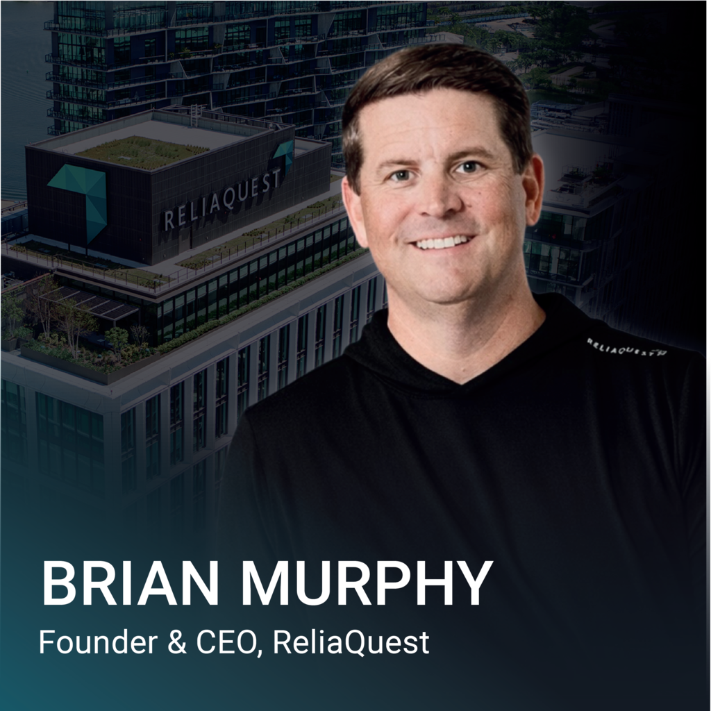 Man standing in front of building with "ReliaQuest" logo. He is wearing a black shirt and smiling.