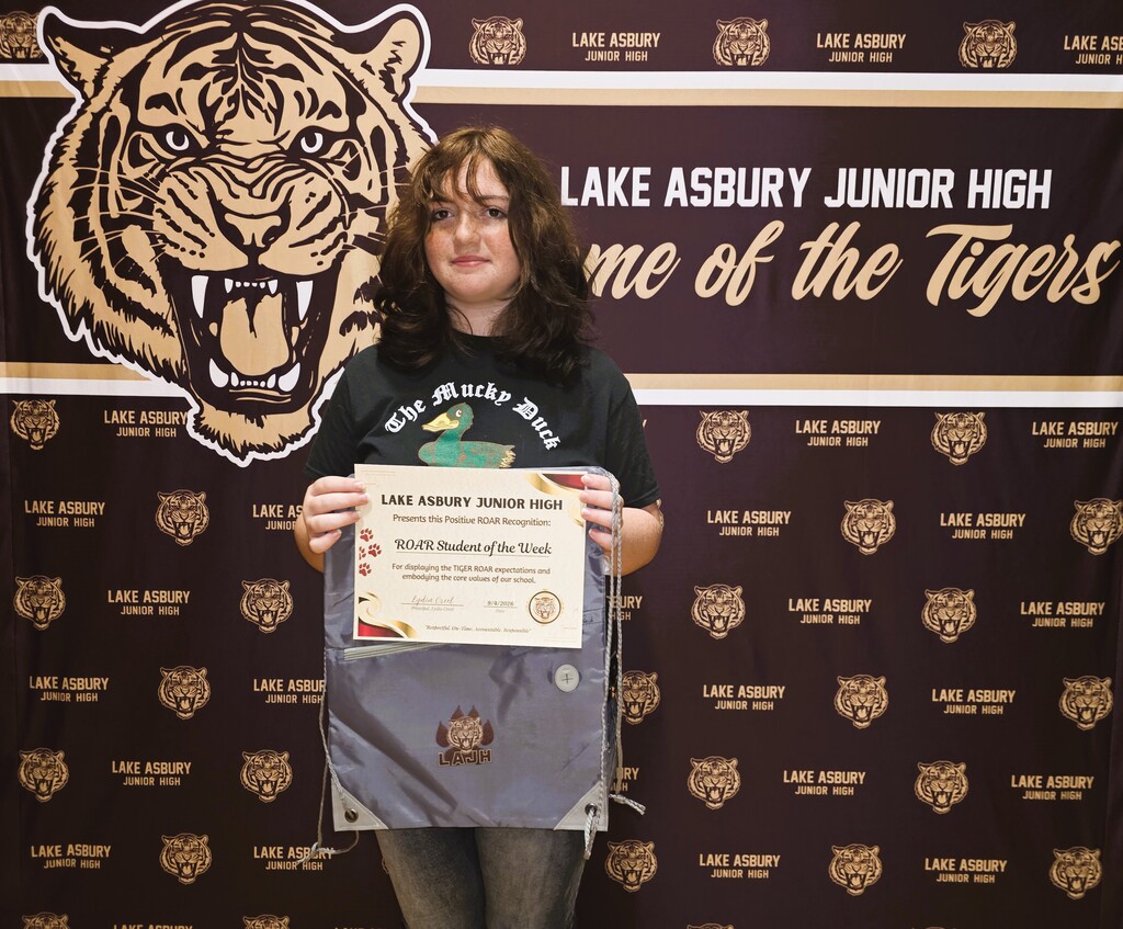 A young woman holding a certificate stands before a backdrop with tiger images and the name of the school.