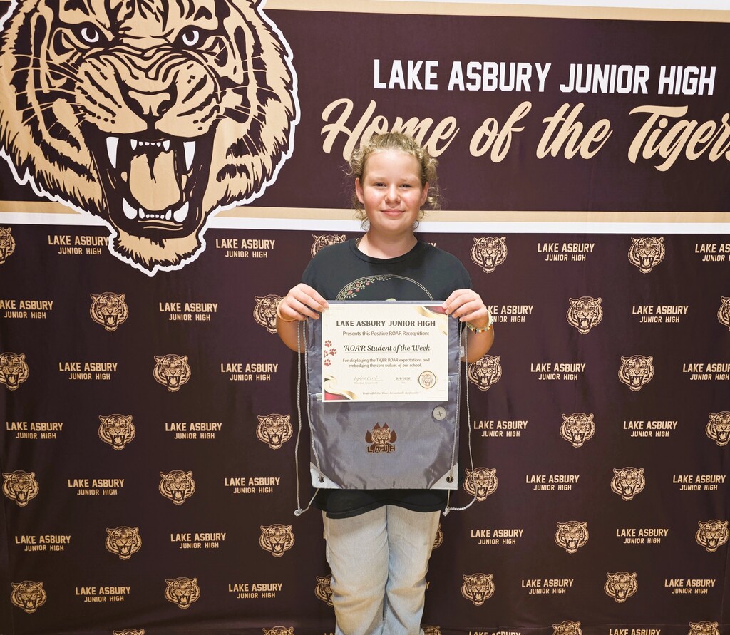 A student holds a certificate in front of a wall with a tiger design and "Lake Asbury Junior High" text.