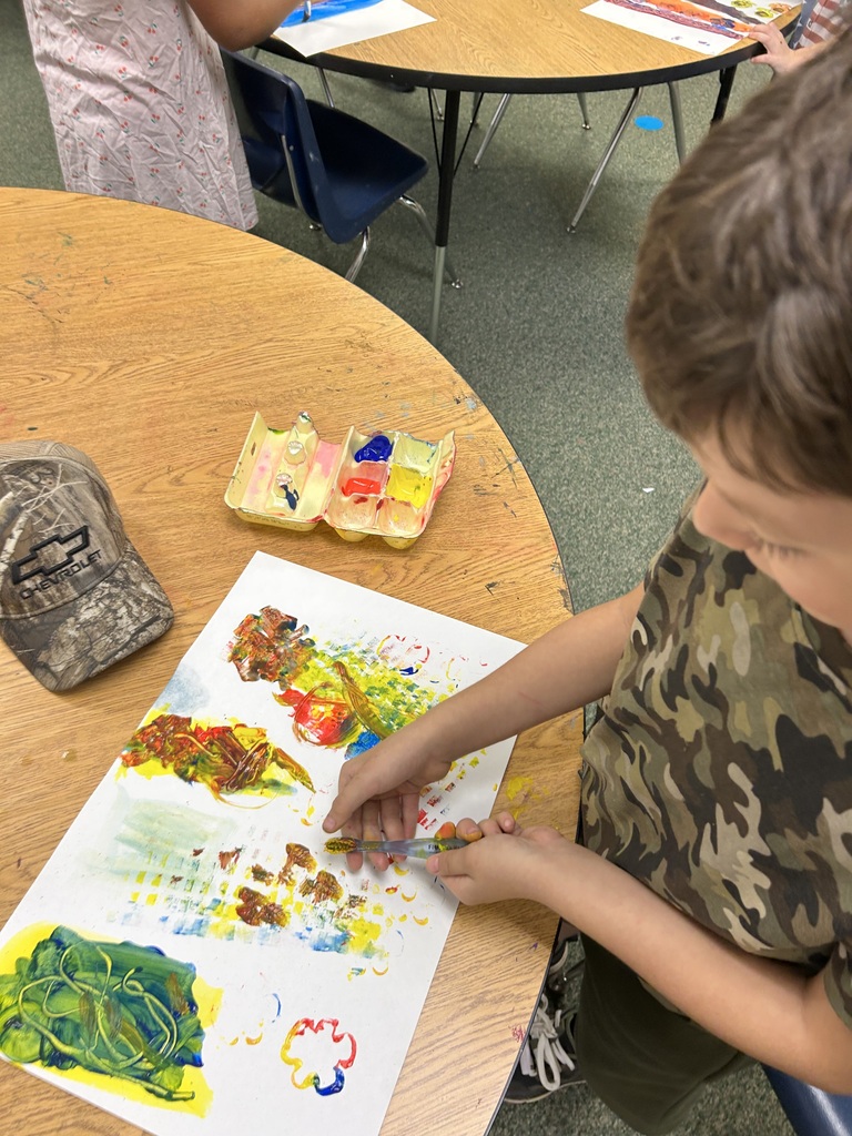 A child uses paintbrushes to paint on a canvas. There is a hat, paint palette, and other art supplies on the table.